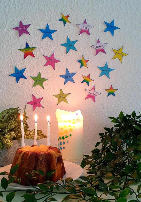 Bild vergrößern: Vor einer Wand, auf der sich Sterne mit Kindernamen darauf befinden, steht auf einem Tisch ein Geburtstagskuchen mit brennenden Kerzen.